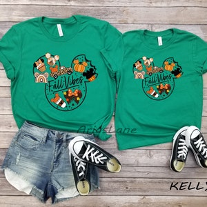 May include: Two green t-shirts featuring a Mickey Mouse head design with autumnal elements such as pumpkins, coffee cups, and a car. The text "Fall Vibes" is printed on the shirts.