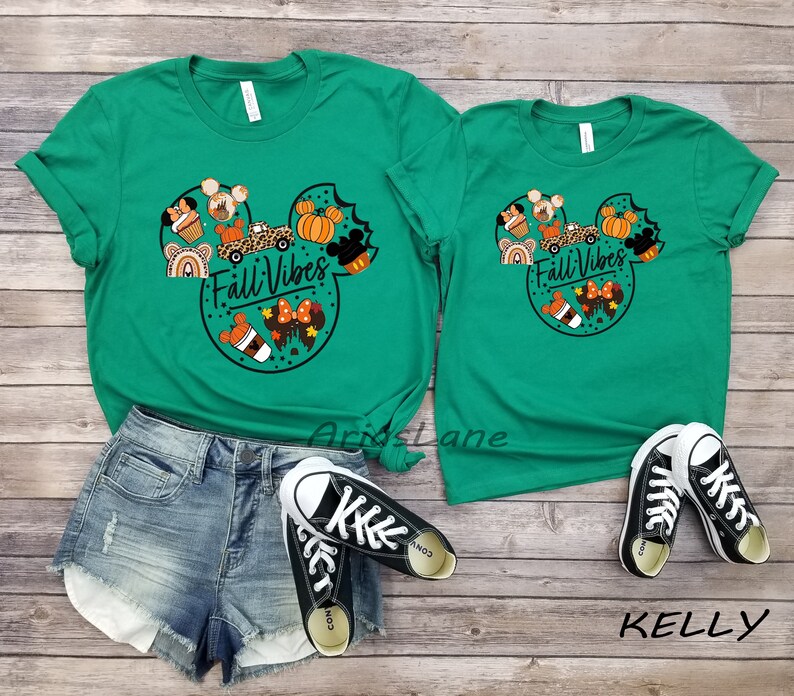 May include: Two green t-shirts featuring a Mickey Mouse head design with autumnal elements such as pumpkins, coffee cups, and a car. The text "Fall Vibes" is printed on the shirts.