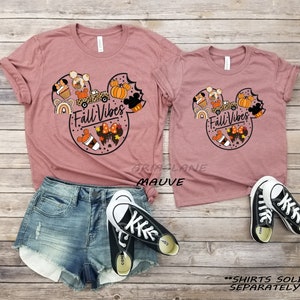 May include: Two mauve t-shirts featuring a Mickey Mouse head design with autumnal elements such as pumpkins, coffee, and a rainbow. The text "Fall Vibes" is inside the Mickey Mouse head. The shirts are sold separately.