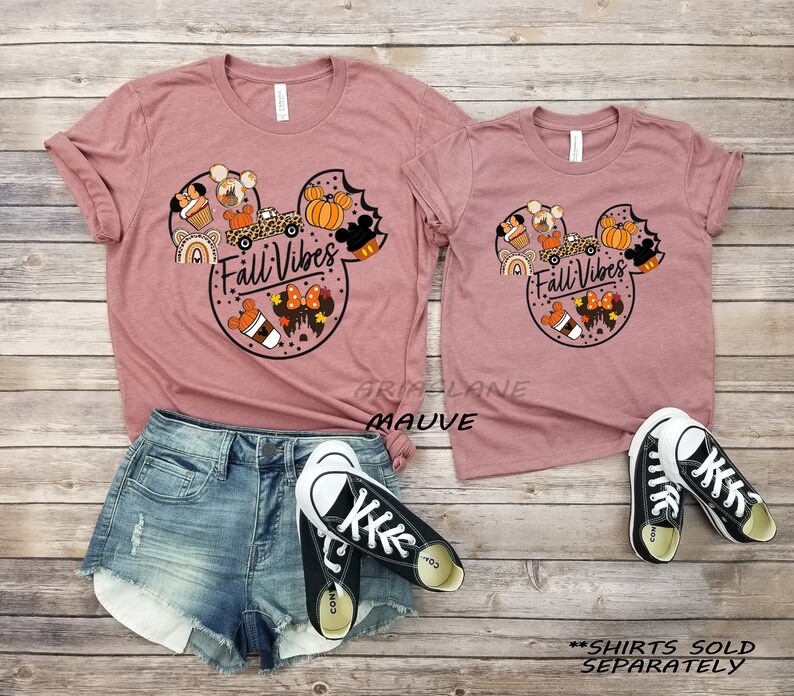 May include: Two mauve t-shirts featuring a Mickey Mouse head design with autumnal elements such as pumpkins, coffee, and a rainbow. The text "Fall Vibes" is inside the Mickey Mouse head. The shirts are sold separately.