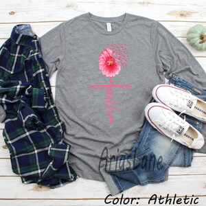 May include: A long-sleeved t-shirt in athletic grey with a pink flower graphic and the word "Faith" in pink lettering. The shirt is shown on a model wearing blue jeans and white trainers.