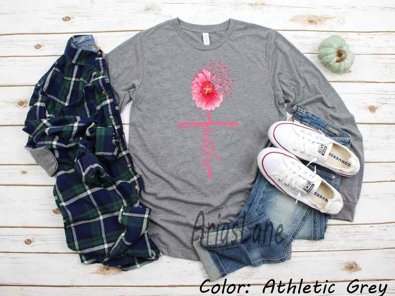 May include: A long-sleeved t-shirt in athletic grey with a pink flower graphic and the word "Faith" in pink lettering. The shirt is shown on a model wearing blue jeans and white trainers.