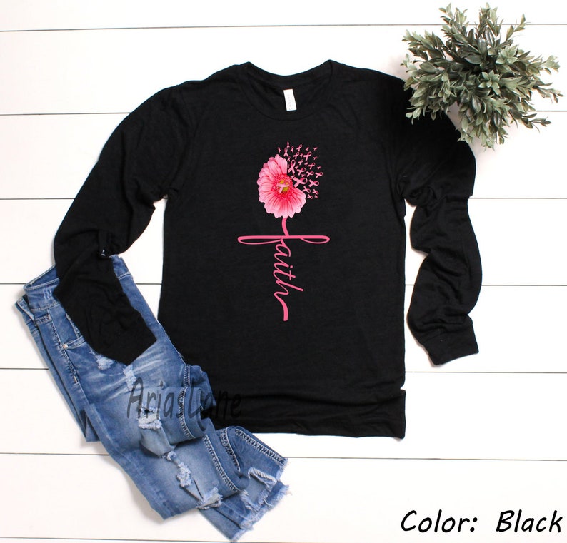 May include: Black long-sleeved shirt featuring a pink flower design and the word "faith" written vertically. The flower is made up of pink ribbons.