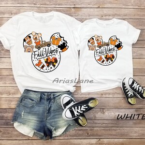 May include: Two white t-shirts with a Mickey Mouse head design featuring autumn-themed elements like pumpkins, coffee, and a truck. The text "Fall Vibes" is written inside the Mickey Mouse head. The shirts are laid out on a wooden surface with a pair of black trainers.