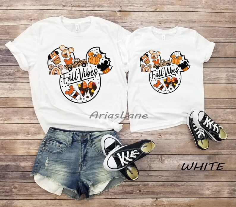 May include: Two white t-shirts with a Mickey Mouse head design featuring autumn-themed elements like pumpkins, coffee, and a truck. The text "Fall Vibes" is written inside the Mickey Mouse head. The shirts are laid out on a wooden surface with a pair of black trainers.