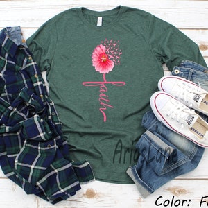 May include: A green long-sleeved t-shirt featuring a pink flower with the word "faith" written vertically. The flower has pink ribbons that appear to be blowing in the wind. The shirt is displayed on a white wooden background with a plaid shirt, blue jeans, and white trainers.