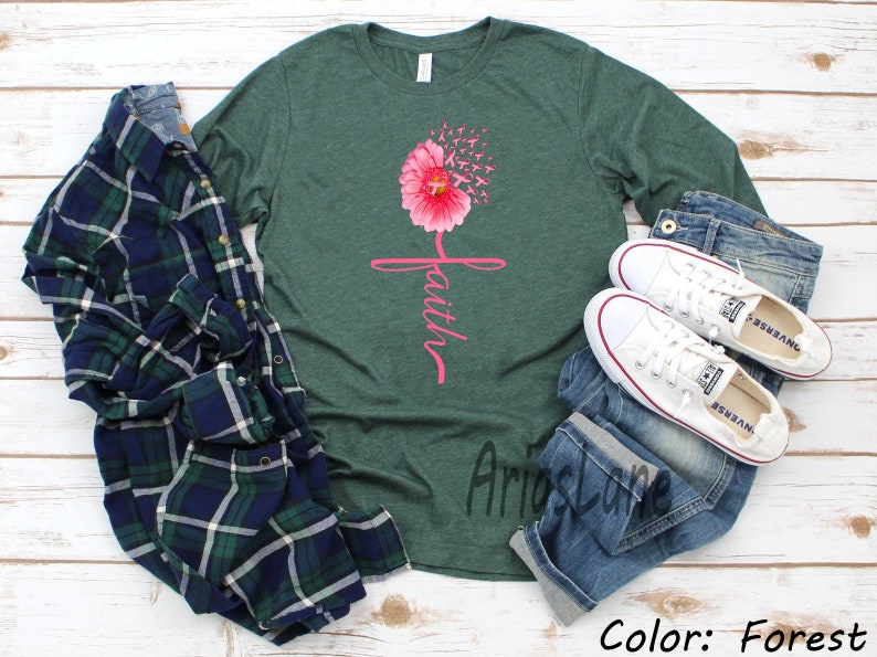 May include: A green long-sleeved t-shirt featuring a pink flower with the word "faith" written vertically. The flower has pink ribbons that appear to be blowing in the wind. The shirt is displayed on a white wooden background with a plaid shirt, blue jeans, and white trainers.