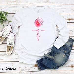 May include: White long-sleeved t-shirt featuring a pink flower design and the word "faith" written vertically. The flower is made up of pink ribbon symbols.