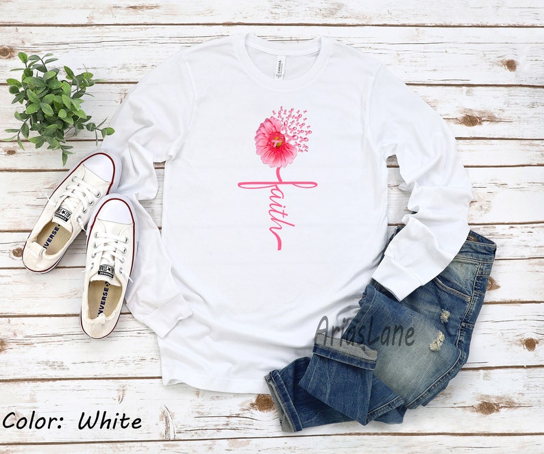 May include: White long-sleeved t-shirt featuring a pink flower design and the word "faith" written vertically. The flower is made up of pink ribbon symbols.