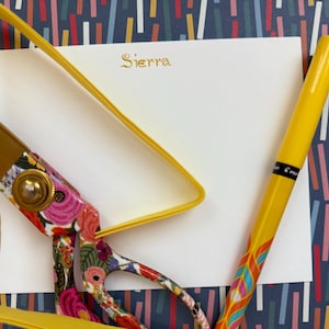 May include: A white card with the name "Sierra" written in gold script on a blue and white patterned background. A yellow pen and a pair of floral scissors are on the card.