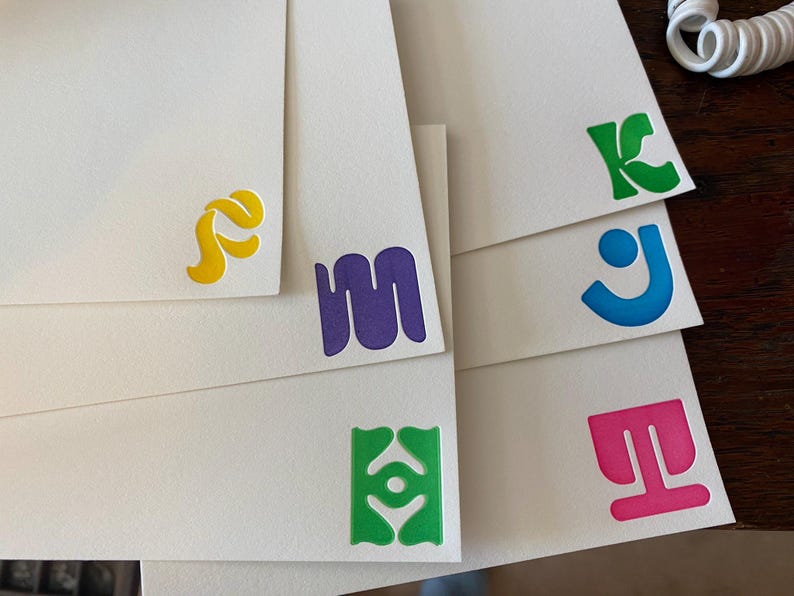 May include: A set of six blank cards with colourful, abstract designs. The designs include a yellow swirl, a purple squiggle, a green eye-like shape, a pink T-shaped design, a blue J-shaped design, and a green K-shaped design.