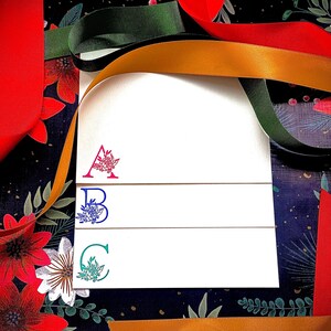 May include: Three white cards with a floral design and the letters A, B, and C in different colours. The cards are on a dark background with red and gold ribbon.