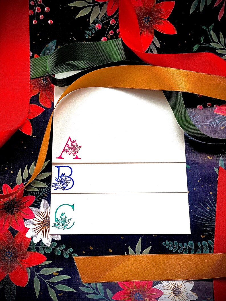 May include: Three white cards with a floral design and the letters A, B, and C in different colours. The cards are on a dark background with red and gold ribbon.