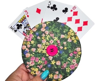 May include: A round, fabric coin purse with a floral print in shades of pink, yellow, and green. A pink button is in the center of the purse. Playing cards are fanned out behind the purse, showing the King of Clubs, 3 of Hearts, 5 of Clubs, and 10 of Diamonds.