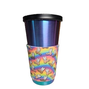 Puede incluir: Un vaso de acero inoxidable con tapa negra y funda colorida. La funda presenta un patrón repetido de penes alados sobre un fondo arcoíris. El vaso tiene un acabado metálico azul y morado.