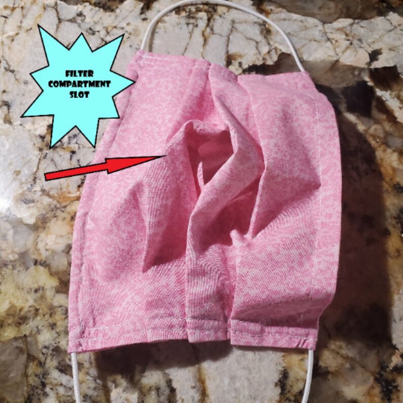May include: Pink fabric face mask with a filter compartment slot. The mask is pleated and has white stitching.