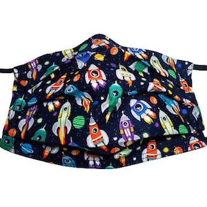 May include: A navy blue face mask with a repeating pattern of colorful rockets, planets, and stars. The rockets are in various colors, including red, orange, green, and purple. The mask has black elastic ear loops.