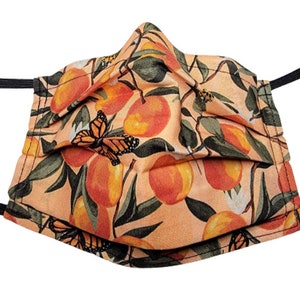 Könnte beinhalten: Eine pfirsichfarbene Gesichtsmaske mit einem sich wiederholenden Muster aus orangenen Früchten und Monarchfaltern. Die Maske hat schwarze elastische Ohrschlaufen.