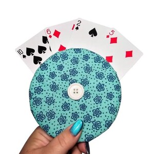 May include: A teal blue fabric coin purse with a white button and a floral pattern. The purse is holding a stack of playing cards, including the 10 of spades, 6 of diamonds, 2 of spades, and 5 of diamonds.
