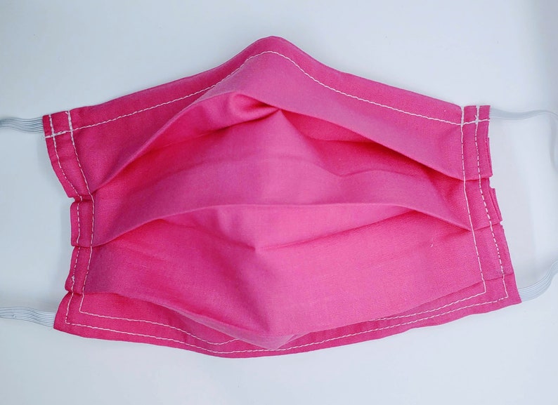 May include: A pink face mask with white stitching. The mask is pleated and has elastic ear loops.