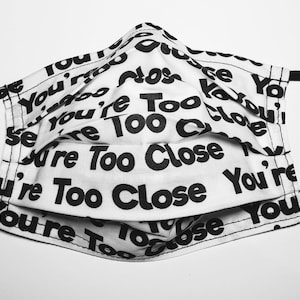 Puede incluir: Una mascarilla facial blanca con correas elásticas negras y la frase repetida "You're Too Close" impresa en la tela.