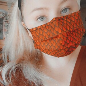 May include: A person wearing a brown v-neck shirt and a face mask with an orange and brown dragon scale pattern.