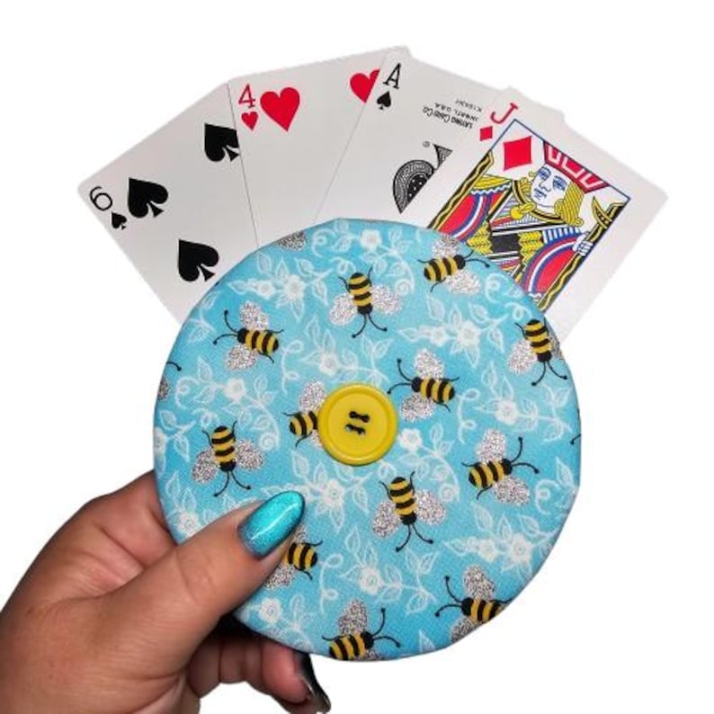 Puede incluir: Una cartera de monedas de tela azul y blanca con un bot&oacute;n amarillo y un patr&oacute;n de abejas. La cartera est&aacute; decorada con purpurina plateada.