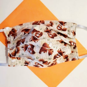 May include: A beige face mask with a repeating pattern of brown foxes. The mask has elastic ear loops and is pleated for a comfortable fit.