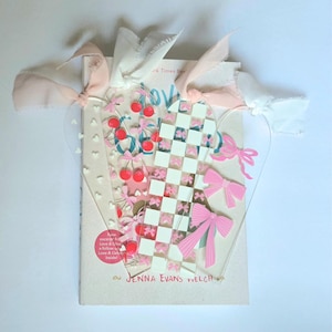 May include: A decorative bookmark with a clear heart-shaped design, featuring pink bows, cherries, and a checkered pattern. The bookmark is attached to a book with the title "Love & Live" and the author's name "Jenna Evans Welch". Pink and white ribbons are tied to the top.
