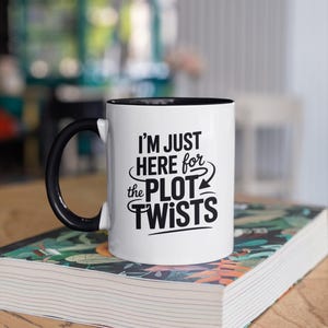 May include: White ceramic mug with a black interior and handle. The mug features the text "I'M JUST HERE for the PLOT TWISTS" in black lettering. The mug is sitting on a book with a colorful cover.