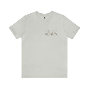 May include: A light gray t-shirt with short sleeves. The shirt features the word "Grassroots" in a script font on the front, along with a small graphic. A small logo is visible near the neckline.