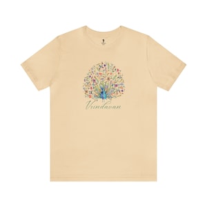 Könnte beinhalten: Ein beiges T-Shirt mit einem Pfauen-Design in einem Kreis aus Blumen. Der Pfau ist blau und grün mit einem langen Schwanz. Der Text "Vrindavan" ist in Grün unter dem Pfau geschrieben.