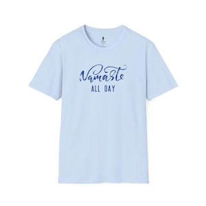 May include: A light blue t-shirt with the words "Namaste ALL DAY" in a dark blue, stylized script. The shirt has short sleeves and a crew neck. The text is centered on the front of the shirt.