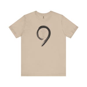 Può includere: Una maglietta beige con un designo a pennellate nere del numero nove. Il numero nove è formato da un cerchio con una linea che si estende verso il basso dal cerchio.