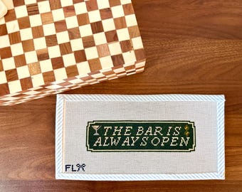 May include: A needlepoint sign with the text "THE BAR IS ALWAYS OPEN" in green and white. The sign has a light blue border and the letters "FLX" in the lower left corner. A wooden box with a checkerboard pattern is in the background.