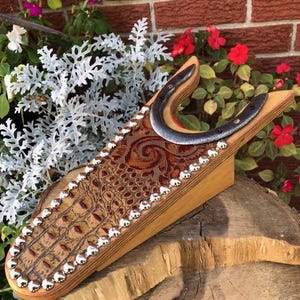 Peut inclure: Un tire-bottes en bois avec une incrustation en cuir marron ornée d'un motif crocodile et de clous décoratifs argentés. Un fer à cheval en métal est fixé sur le dessus. Le tire-bottes est posé sur une surface en bois, avec des fleurs en arrière-plan.
