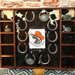 Op de afbeelding: Een houten vitrinekast met een koeienhuid achtergrond met tien metalen hoefijzers in een cirkel. Een centraal bord toont een oranje cowboyhoed, een slangenkop en de tekst Copperhead Dave. Er zijn diverse metalen riemgespen in verwerkt.