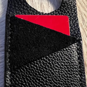Könnte beinhalten: Schwarzer Lederkartenhalter mit strukturierter Oberfläche. Verfügt über eine Tasche mit schwarzem Wildlederfutter und einer teilweise sichtbaren roten Karte. Inklusive Schlaufe. Ein stilvolles Accessoire für den täglichen Gebrauch.