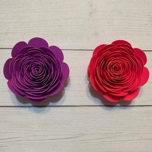 May include: Two paper flowers, one purple and one red, are arranged on a white wooden surface.