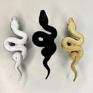 May include: Three snake-shaped cabinet knobs arranged vertically. The left knob is silver with a textured surface, the center knob is matte black, and the right knob is gold with a textured finish. Each knob features a coiled snake design.