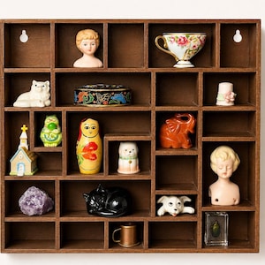 May include: A brown wooden display cabinet with numerous compartments, showcasing a collection of vintage items. These include a porcelain doll bust, a floral teacup, a cat figurine, a Russian doll, and a small church ornament.