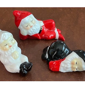 May include: Three ceramic figurines of sleeping figures with white beards. One figure wears a red suit and hat, another is entirely white with black boots, and the third wears a black suit and hat with red accents. All have closed eyes and rosy cheeks.