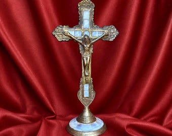 May include: A detailed brass crucifix with a bronze figure of Jesus. The cross features ornate detailing and light blue accents. The base is round with a light blue and white mosaic design. The background is a deep red fabric.