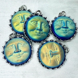 May include: Five iridescent moon face pendants with silver-colored metal trim. The faces have a smiling expression and are in shades of blue, green, and gold. Each pendant has a small loop at the top for hanging.