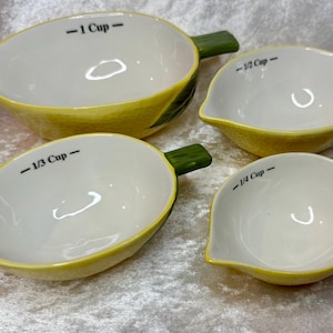 Può includere: Un set di quattro misurini in ceramica a forma di limone con gambi verdi. I misurini sono etichettati 1 Cup, 1/2 Cup, 1/3 Cup e 1/4 Cup in nero. Ogni misurino ha un esterno giallo, un interno bianco e un beccuccio appuntito per versare.