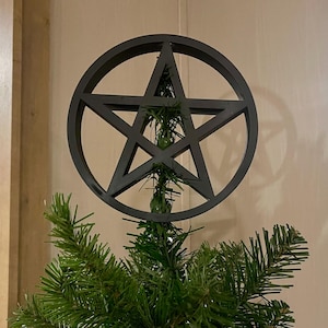 Può includere: Un ornamento a pentagramma nero opaco caratterizzato da una stella a cinque punte racchiusa in una cornice circolare. L'oggetto è posizionato sulla cima di un albero di pino artificiale verde, con i rami che passano attraverso il centro.