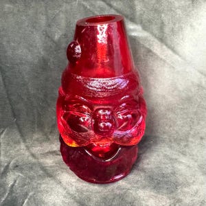 Könnte beinhalten: Eine durchscheinende rote Glasfigur in Form eines stilisierten Weihnachtsmannkopfes. Das Stück zeigt eine spitze Mütze mit einem kreisförmigen Bommel-Detail, einen breiten Schnurrbart und eine abgerundete Nase. Das Glas hat eine glatte Oberfläche.