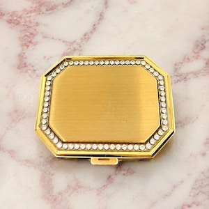 May include: A rectangular gold-toned compact mirror with beveled edges and a brushed metal finish. The perimeter of the lid is framed by a single row of clear, sparkling rhinestones set into the gold metal border, featuring a small latch.