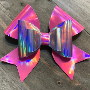 Pode incluir: Um grande laço rosa iridescente com acabamento holográfico. O laço é feito de um material brilhante e tem uma forma tridimensional.