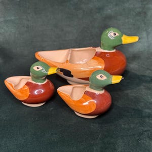 Peut inclure: Un ensemble de trois bols en céramique en forme de canard colvert avec des têtes vertes, des becs jaunes et des corps marron et orange. Le plus grand bol a un intérieur divisé, tandis que les deux plus petits ont des compartiments uniques.